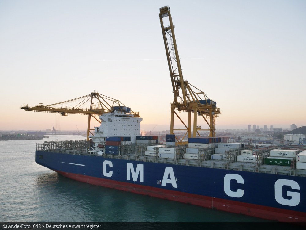 Dieses Foto zeigt einen Teil eines halbvoll beladenes Containerschiff im Hafen von Barcelona (Spanien). Im Hintergrund sind zwei große Verladekräne zu sehen. Auf der Seitenwand des Containerschiff findet sich die Aufschrift CMA CGM als Hinweis auf die Reederei.