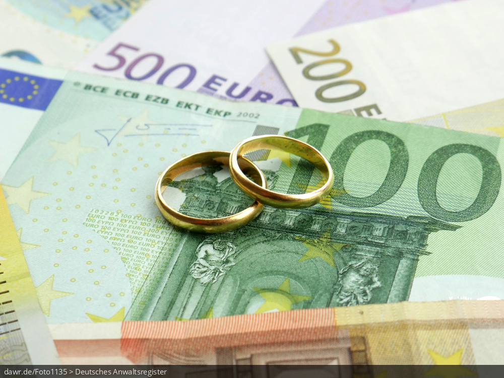 Dieses Foto zeigt Geldscheine, die auf einer Fläche verteilt wurden. Auf den Banknoten liegen zwei Eheringe. Dieses Bild eignet sich gut als symbolische Darstellung für alle Themen rund um Geld und Ehe.