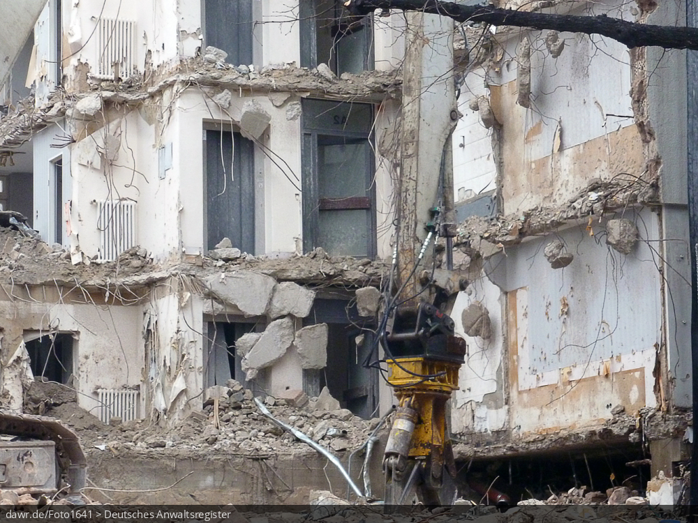 Dieses Foto zeigt eine Baustelle mit einem teilweise abgebrochenen Haus. Im Vordergrund ist ein Bagger mit einer Abbruchzange zu sehen, wie sie zum Durchtrennen der Stahlarmierungen beim Abbruch von Gebäuden aus Stahlbeton verwendet. Dieses Bild ist gut als symbolhafte Darstellung für Streitigkeiten in Zusammenhang mit Abrissarbeiten (oder allgemein Bauarbeiten) geeignet.