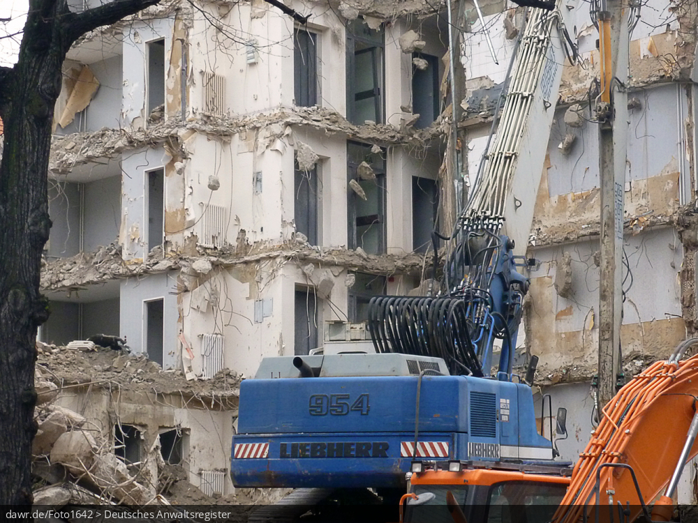 Dieses Foto zeigt eine Baustelle mit einem teilweise abgebrochenen Haus. Im Vordergrund ist ein Bagger mit einer Abbruchzange zu sehen, wie sie zum Durchtrennen der Stahlarmierungen beim Abbruch von Gebäuden aus Stahlbeton verwendet. Dieses Bild ist gut als symbolhafte Darstellung für Streitigkeiten in Zusammenhang mit Abrissarbeiten (oder allgemein Bauarbeiten) geeignet.