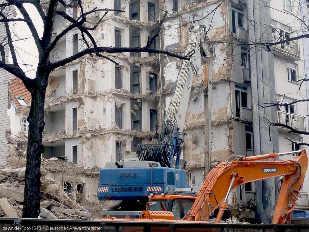 Dieses Foto zeigt eine Baustelle mit einem teilweise abgebrochenen Haus. Im Vordergrund ist ein Bagger mit einer Abbruchzange zu sehen, wie sie zum Durchtrennen der Stahlarmierungen beim Abbruch von Gebäuden aus Stahlbeton verwendet. Dieses Bild ist gut als symbolhafte Darstellung für Streitigkeiten in Zusammenhang mit Abrissarbeiten (oder allgemein Bauarbeiten) geeignet.