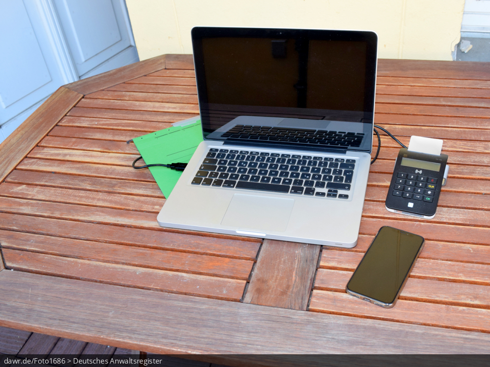 Dieses Foto zeigt einen Holztisch auf dem ein Notebook, ein Smartphone und Hängehefter liegen. An den Computer ist ein Kartenlesegerät angeschlossen, mit dem auch Smartcards gelesen werden können, wie sie bspw. beim besonderen elektronischen Anwaltspostfach (beA) genutzt werden. Dieses Bild eignet sich besonders gut als symbolische Darstellung für das Thema Home-Office, wie es im Zusammenhang mit der Corona-Pandemie sehr populär geworden ist. Durch die Darstellung des Kartenlesers ist auch eine Verwendung im Zusammenhang mit dem besonderen elektronischen Anwaltspostfach (beA) denkbar.