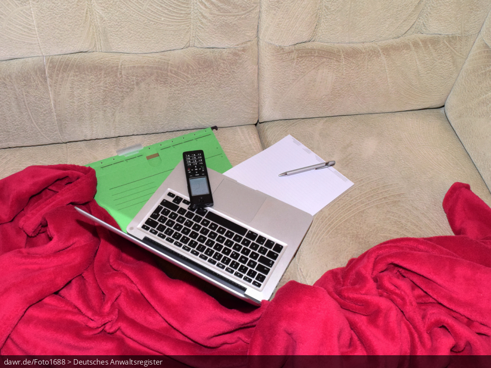 Dieses Foto zeigt ein Sofa auf dem ein Notebook, ein Telefon, ein Block (mit Kugelschreiber) und ein Hängehefter zu sehen sind. Umrahmt wird die Szenerie durch eine rote Decke, wie sie viele auf ihrer Couch liegen haben. Dieses Bild eignet sich gut als symbolische Darstellung für das Thema Home-Office, wie es im Zusammenhang mit der Corona-Pandemie sehr populär geworden ist.