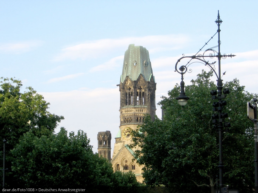 Dieses Foto zeigt die evangelische Kaiser-Wilhelm-Gedächtniskirche in Berlin, fotografiert durch einige Bäume hindurch. Die Kirche wird auch kurz Gedächtniskirche oder im Volksmund „Hohler Zahn“ genannt und liegt zwischen dem Kurfürstendamm und der Tauentzienstraße im Ortsteil Charlottenburg, driekt am Breitscheidplatz.