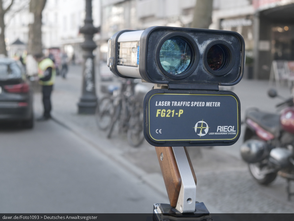 Dieses Foto zeigt ein Lasergeschwindigkeitsmessgerät (gern auch Laserpistole genannt) vom Typ RIEDL FG21-P. Unscharf im Hintergrund ist eine Polizeikontrolle zu sehen.