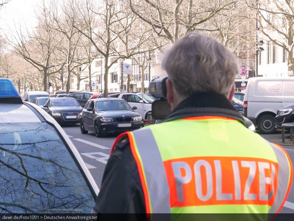 Dieses Foto zeigt einen Polizisten bei einer Geschwindigkeitskontrolle mit einem Lasergeschwindigkeitsmessgerät (gern auch Laserpistole genannt) auf dem Kurfürstendamm in Berlin. Der Beamte trägt eine leuchtend gelb-orange Warnweste mit dem Aufdruck Polizei, während er die Messung mit einem LASER TRAFFIC SPEED METER FG21-P der Firma Riedl durchführt.