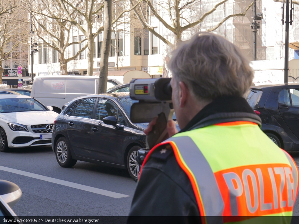 Dieses Foto zeigt einen Polizisten bei einer Geschwindigkeitskontrolle mit einem Lasergeschwindigkeitsmessgerät (gern auch Laserpistole genannt) auf dem Kurfürstendamm in Berlin. Der Beamte trägt eine leuchtend gelb-orange Warnweste mit dem Aufdruck Polizei, während er die Messung mit einem LASER TRAFFIC SPEED METER FG21-P der Firma Riedl durchführt.