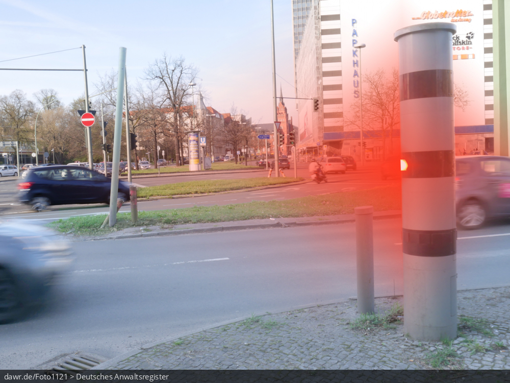 Dieses Foto zeigt eine große Straßenkreuzung, an der eine Geschwindigkeitsüberwachungsanlage vom Typ Vitronic Poliscan speed aufgestellt wurde, welche gerade ein Auto blitzt. Auch wenn hier ein spezieller Typ von Geschwindigkeitsüberwachungsanlage dargestellt wird, ist es möglich dieses Bild als symbolische Darstellung für Themen in Zusammenhang mit Geschwindigkeitsverstößen und ihren Konsequenzen zu verwenden.