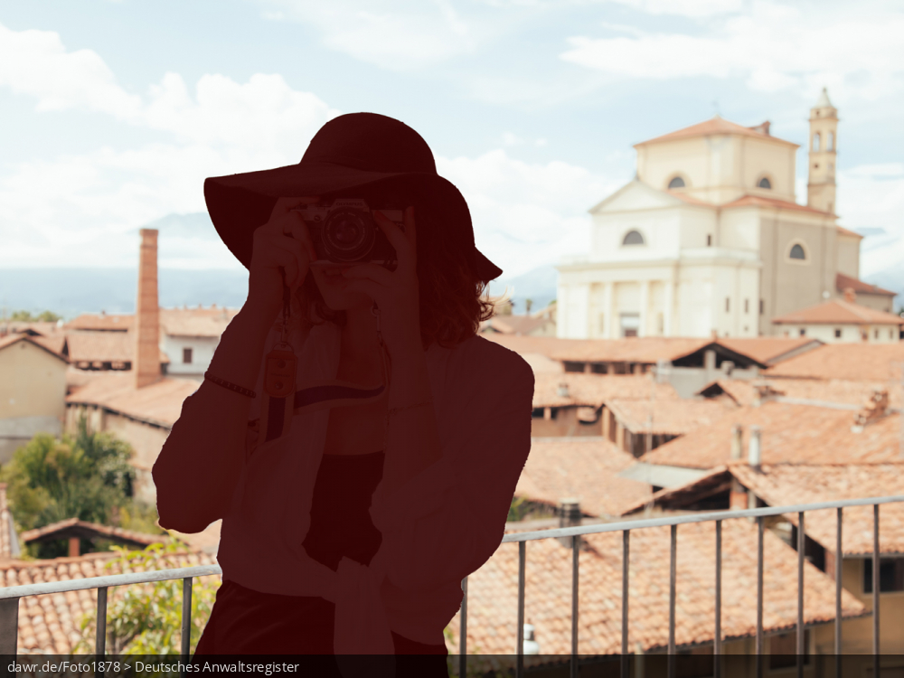 Dieses Foto zeigt eine mediterrane Stadt und davor eine Frau, die Urlaubsfotos macht. Die Frau ist allerdings nur als Silhouette dargestellt, ganz so als wäre sie ausradiert worden. Diese Darstellung eignet sich wegen dem Ausradieren gut für das Thema Reiserücktritt.
