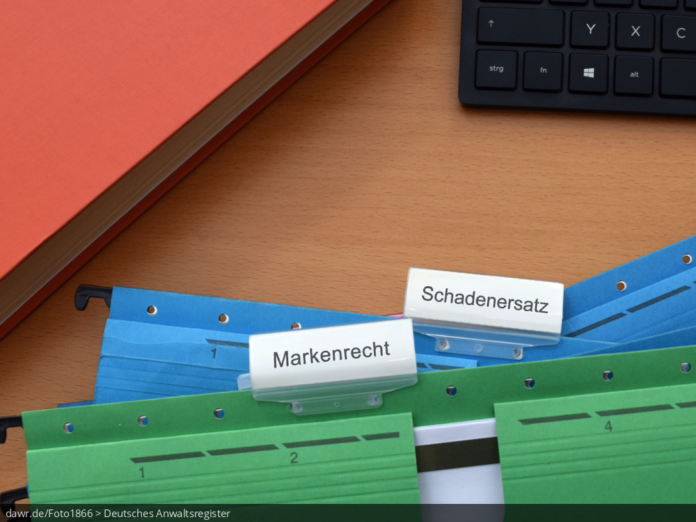 Dieses Foto zeigt ein Aktenstapel, welcher neben einem Fachbuch und einer Tastatur liegt. Hierbei sind die beiden obersten Akten mit „Markenrecht“ und „Schadenersatz“ beschriftet. Diese Darstellung eignet sich gut für Themen im Zusammenhang mit Markenrechtsverletzungen.