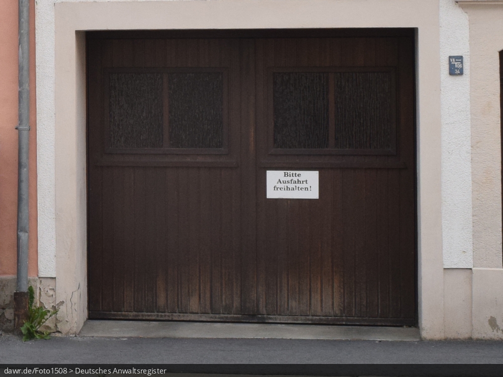 Das Foto zeigt das Holztor einer Garage mit einem Hinweisschild mit der Aufschrift „Bitte Ausfahrt freihalten!“. Viele Grundstückseigentümer bringen solche Schilder an ihren Ausfahrten an, um das Zuparken dieser zu vermeiden. Dieses Bild eignet sich gut als symbolische Darstellung für das Thema Parken in der Stadt.
