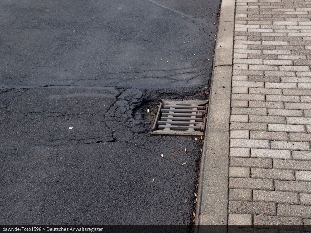 Dieses Foto zeigt eine Hauptverkehrsstraße in einer deutschen Stadt. Man sieht deutliche Risse im Asphalt und am Rande eines Gullis ein ausgeprägtes Schlagloch. Dieses Bild ist gut als symbolhafte Darstellung für das Auftreten von Straßenschäden und die damit einhergehenden rechtlichen Fragen, bspw. im Schadenersatzrecht.