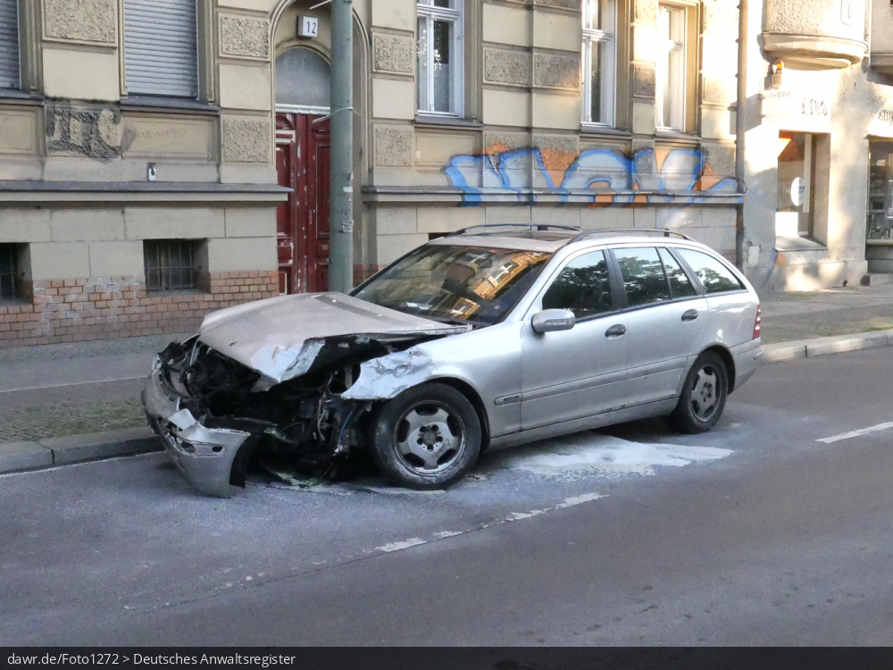 Dieses Foto zeigt ein bei einem Unfall stark beschädigtes Auto, wobei auf der Straße noch Reste von Öl-Bindemittel zu sehen sind. Dieses Bild eignet sich gut als symbolische Darstellung für die Themen Verkehrsunfall und Folgeschäden.