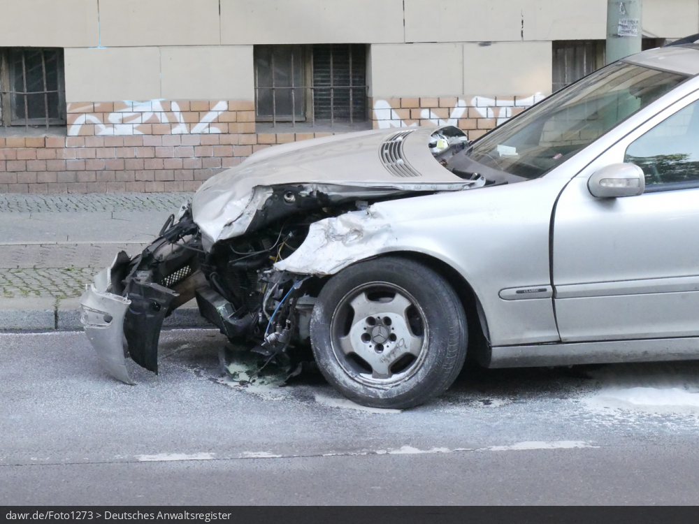 Dieses Foto zeigt ein bei einem Unfall stark beschädigtes Auto, wobei auf der Straße noch Reste von Öl-Bindemittel zu sehen sind. Dieses Bild eignet sich gut als symbolische Darstellung für die Themen Verkehrsunfall und Folgeschäden.