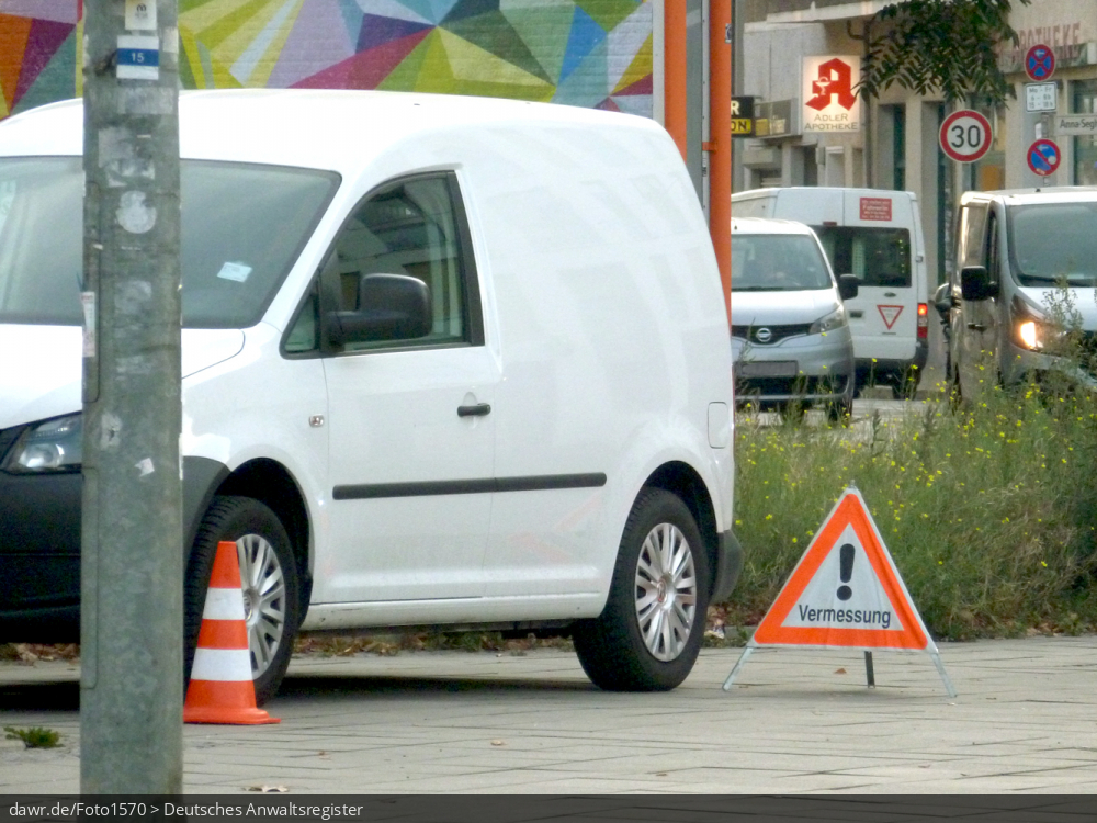 Dieses Foto zeigt das Fahrzeug eines Vermessungsbüros, wie es während eines Einsatzes im öffentlichen Straßenraum abgestellt wurde. Davor sieht man Warnschild, wie es aufgestellt wird, um Passanten vor Beeinträchtigung durch den Vermessungseinsatz zu warnen. Dieses Bild eignent sich als symbolische Darstellung für alle rechtlichen Fragen im Zusammenhang mit Vermessungen oder allgemein der Tätigkeit von Vermessungsingeneuren.