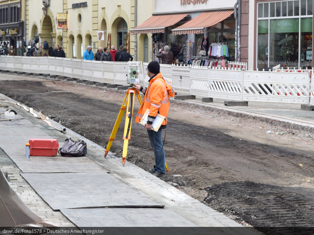 Dieses Foto zeigt den Mitarbeiter eines Vermessungsbüros, wie er Vermessungsarbeiten auf einer Straßenbaustelle vornimmt. Er nutzt dabei ein Nivelliergerät - ein typisches Arbeitsgerät für Vermessungsingeneure. Dieses Bild eignent sich als symbolische Darstellung für alle rechtlichen Fragen im Zusammenhang mit Vermessungen oder allgemein der Tätigkeit von Vermessungsingeneuren.