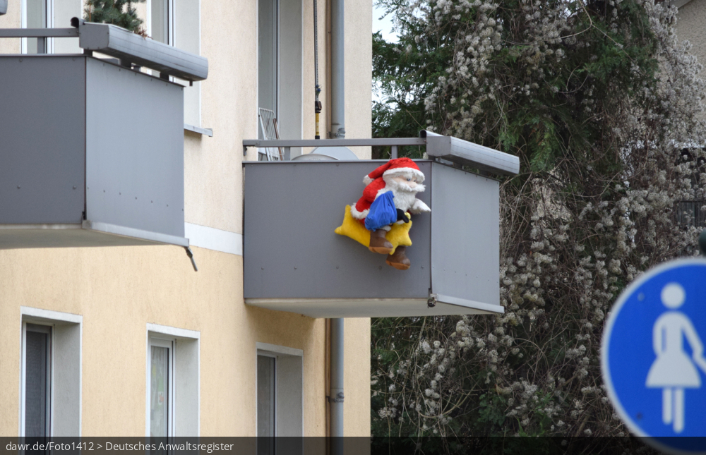 Dieses Foto zeigt einen Weihnachtsmann, welcher am Balkon eines Mehrfamilienhauses hängt. In der heutigen Zeit sind solche Figuren eine gern genutzte Weihnachtsdekoration. Dieses Bild eignet sich gut als symbolische Darstellung für alle Themen rund um Weihnachten und speziell zu rechtlichen Fragen bez. der Weihnachtsdekoration.