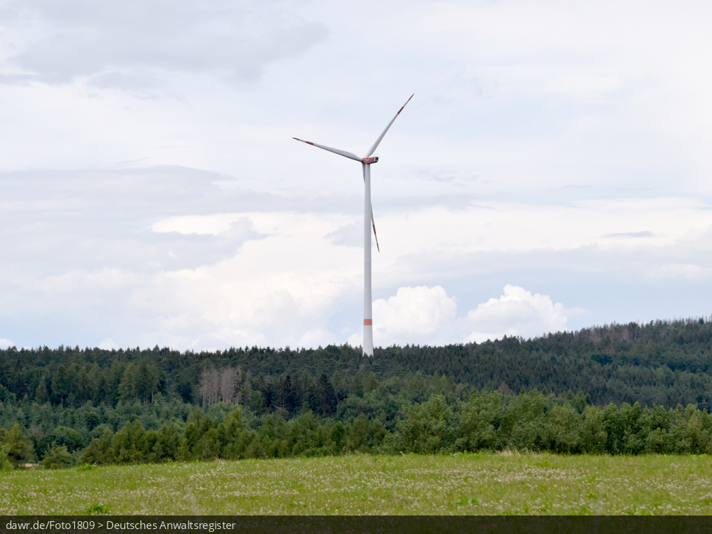Dieses Bild zeigt eine Windkraftanlage, welche in einem umfangreichen Waldgebiet steht, wobei im Vordergrund eine Wiese zu sehen ist. Es ergibt sich so ein passendes Bild für Themen rund um erneuerbare Energien, Windkraft oder allgemein der umweltfreundlichen Stromerzeugung.