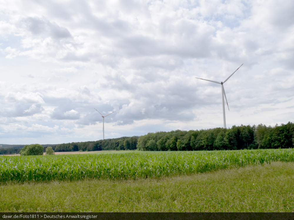 Dieses Bild zeigt zwei Windkraftanlagen, welche in einem umfangreichen Waldgebiet stehen, wobei im Vordergrund ein Feld und eine Wiese zu sehen ist. Es ergibt sich so ein passendes Bild für Themen rund um erneuerbare Energien, Windkraft oder allgemein der umweltfreundlichen Stromerzeugung.