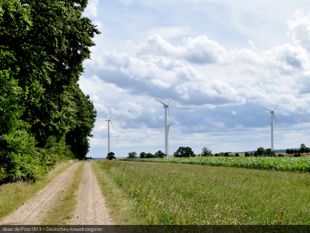 Dieses Bild zeigt mehrere Windkraftanlagen, welche auf Feldern stehen, wobei im Vordergrund auch ein Weg und ein Waldgebiet zusehen sind. Es ergibt sich so ein passendes Bild für Themen rund um erneuerbare Energien, Windkraft oder allgemein der umweltfreundlichen Stromerzeugung.