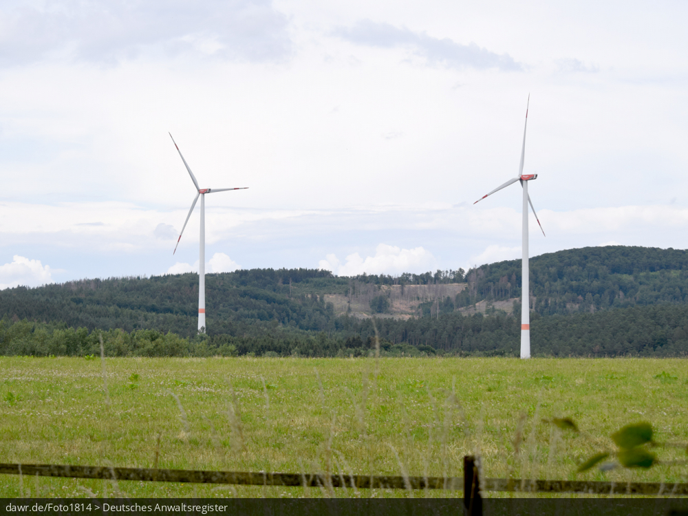 Dieses Bild zeigt zwei Windkraftanlagen auf einer Wiese mit einem umfangreichen Waldgebiet im Hintergrund. Es ergibt sich so ein passendes Bild für Themen rund um erneuerbare Energien, Windkraft oder allgemein der umweltfreundlichen Stromerzeugung.