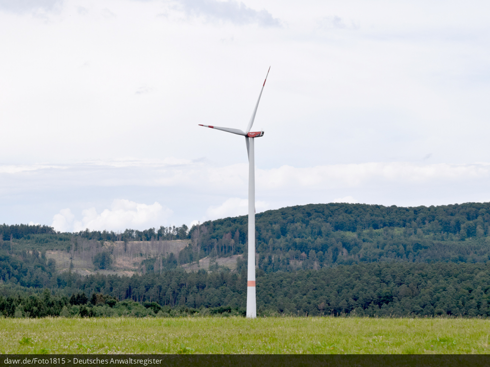 Dieses Bild zeigt eine Windkraftanlage auf einer Wiese mit einem umfangreichen Waldgebiet im Hintergrund. Es ergibt sich so ein passendes Bild für Themen rund um erneuerbare Energien, Windkraft oder allgemein der umweltfreundlichen Stromerzeugung.