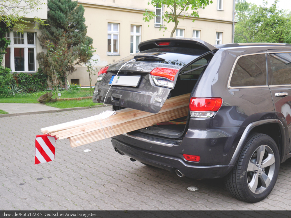Dieses Foto zeigt einen Auto, mit dem lange, schmale Bretter transportiert werden. Die Holzdielen sind so lang, dass hinten aus dem Kofferaum herausragen und mit einer roten Fahne gesichert werden mußten. Dieses Bild eignet sich gut als symbolische Darstellung für das Thema Ladungssicherung beim Auto.