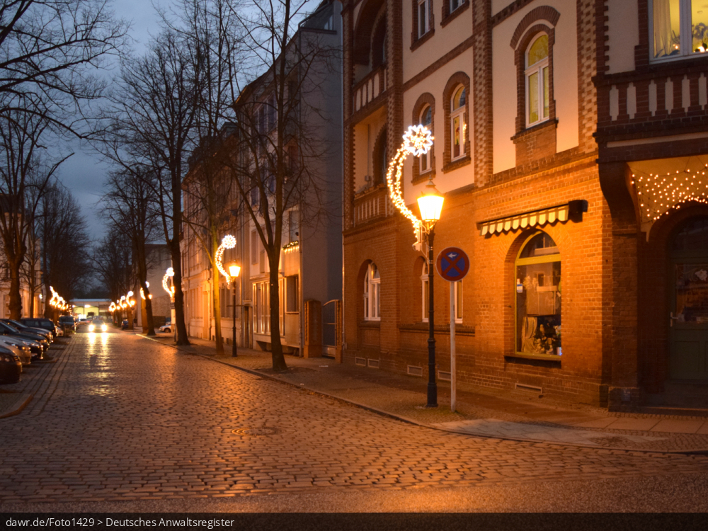 Dieses Foto zeigt eine Nebenstraße in einer Stadt zur Weihnachtszeit. Alle Straßenlaternen wurden mit leuchtenden Sternschnuppen geschmückt, was hier in der Dämmerung besonders gut zum Ausdruck kommt. Diese Abbildung eignet sich gut als symbolische Darstellung für die Themen Weihnachten und Weihnachtsschmuck.