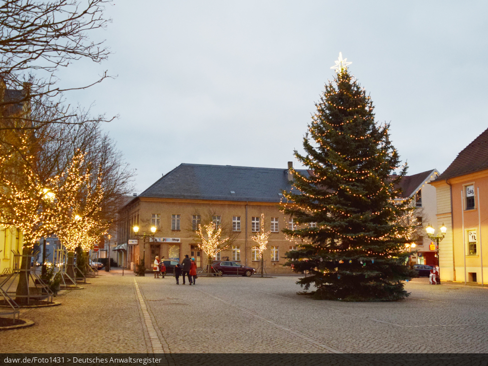 Dieses Foto zeigt einen Platz in einer Stadt zur Weihnachtszeit. Neben einen großen Weihnachtsbaum wurden auf die Straßenbäume mit Lichterketten geschmückt. Diese Abbildung eignet sich gut als symbolische Darstellung für die Themen Weihnachten und Weihnachtsschmuck.