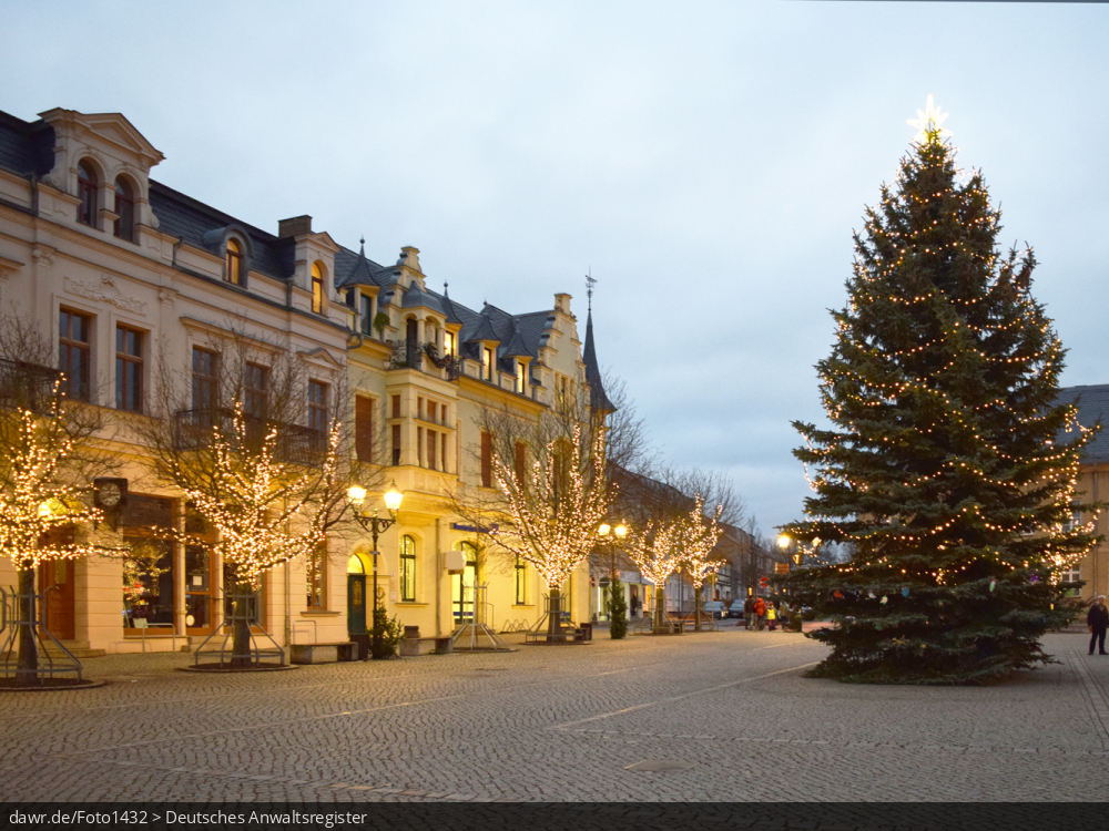 Dieses Foto zeigt einen Platz in einer Stadt zur Weihnachtszeit. Neben einen großen Weihnachtsbaum wurden auf die Straßenbäume mit Lichterketten geschmückt. Diese Abbildung eignet sich gut als symbolische Darstellung für die Themen Weihnachten und Weihnachtsschmuck.