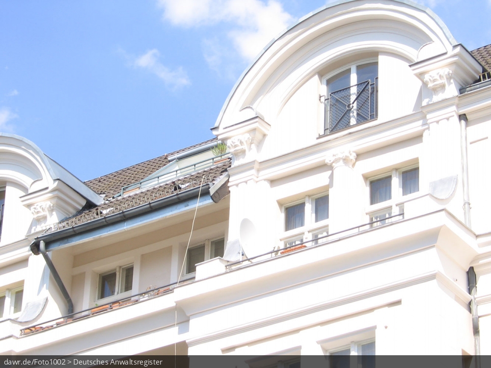 Dieses Foto zeigt die weiße Front eines Mietshauses im Sonnenschein. Es ist der obere Teil des Hauses mit einem langgezogenen Balkon zu sehen.